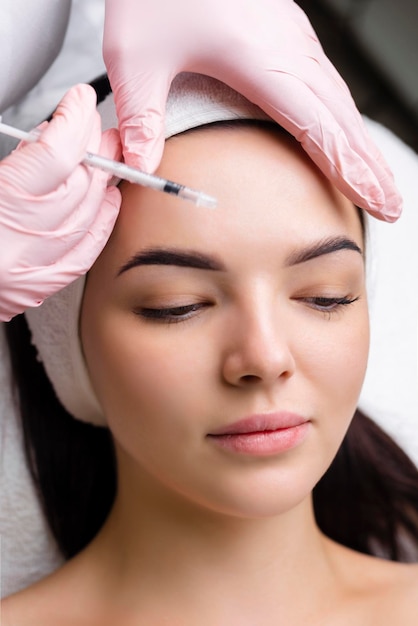 Close up of hands of young cosmetologist injecting botox in female face She is standing and smiling The woman is closed her eyes with relaxation