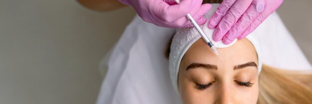 여성 얼굴에 보스를 주사하는 젊은 미용사의 손의 클로즈업 그녀는 서서 미소 짓고 있습니다.