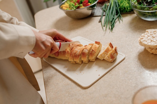 Close up of hands of a young woman cooks in her home kitchen cuts a baguette bread with a knife