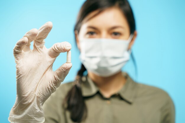 Primo piano delle mani di una giovane donna in una mascherina medica protettiva e guanti che tengono una capsula o una pillola sull'azzurro