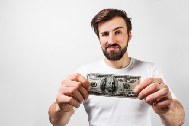 Photo close up of handsome brunette man standing near the white wall and holding a dollar bill. he is showing that he has one hundred dollar bill. guy looks happy. cut view. isolated on white wall.