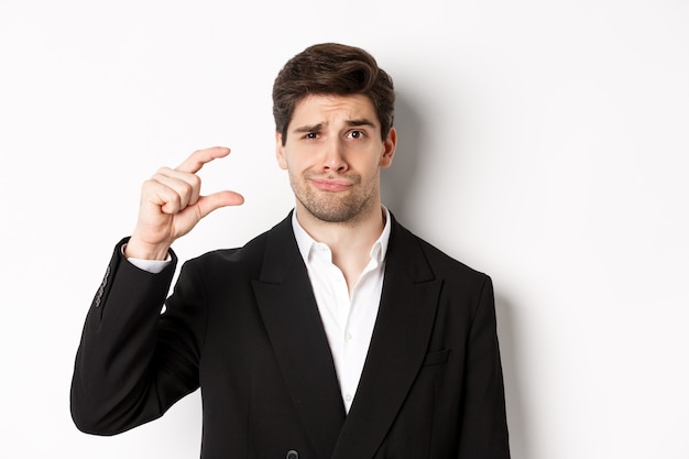 Close-up of handsome businessman in trendy suit, showing something small and looking disappointed, standing against white background