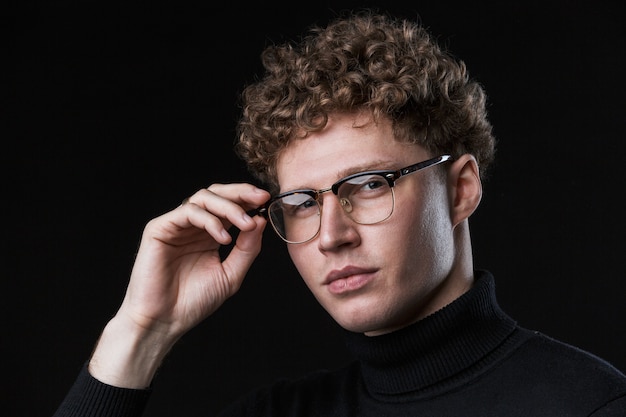 Close up of a handsome confident young curly haired businessman wearing turtleneck standing infront of isolated black wall, posing in glasses