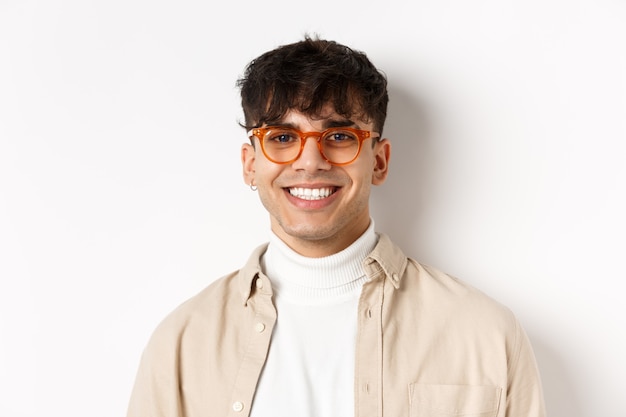 Close-up of handsome hipster guy with natural smile, wearing glasses and earrings, looking happy at camera, standing on white background