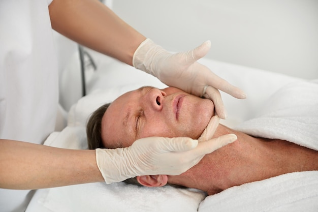 Close-up of handsome mature 50 years old man relaxing on massage table while getting professional facial lifting massage at spa. Anti-aging, rejuvenation, face lifting and skin care procedures concept