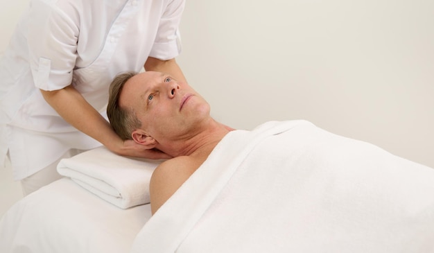 Close-up of handsome mature Caucasian man relaxing on massage table while getting professional facial lifting massage at spa. Anti-aging, rejuvenation, face lifting and skin care procedures concept