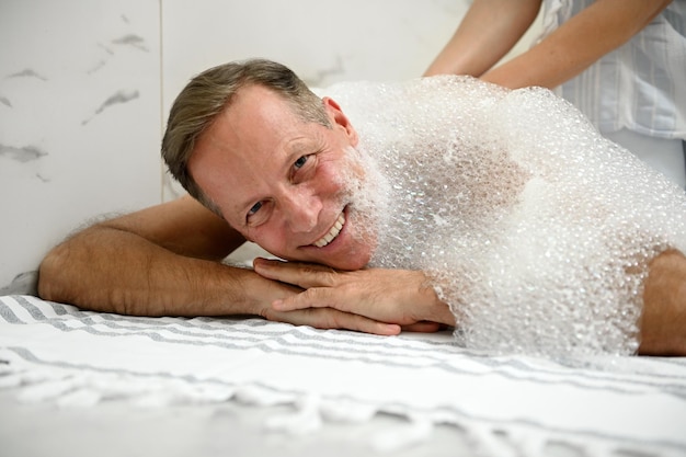 Close-up of a handsome mature European man smiling with beautiful toothy smile, looking at camera while relaxing in Turkish hammam with soap foam on his back