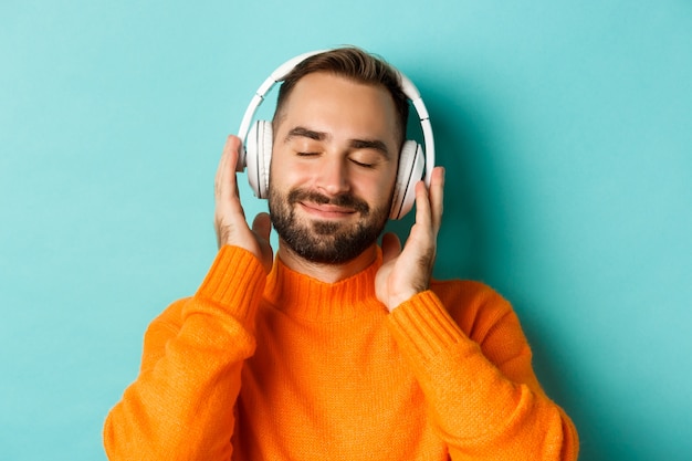 Close-up of handsome modern man listening music in headphones, standing in orange sweater over turquoise background.