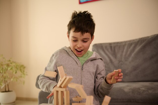 Primo piano di un bel ragazzo europeo in età scolare che gioca a giochi da tavolo e sembra sorpreso quando la sua struttura costruita da blocchi di legno cadono. istruzione per il tempo libero e il concetto di giochi da tavolo