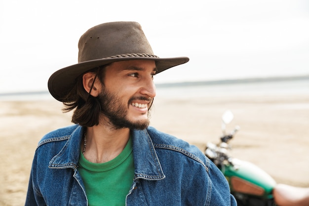 Close up of a handsome smiling young man wearing casual outfit standing at motocycle at the beach