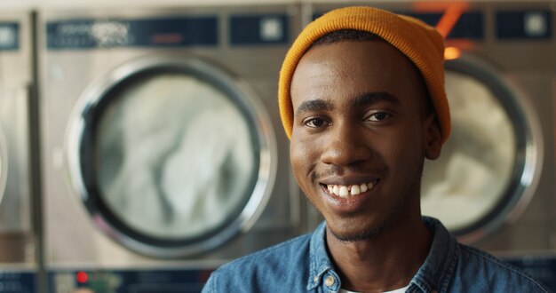 ランドリーサービスルームでカメラに元気よく笑っている黄色い帽子でハンサムな若いアフリカ系アメリカ人のクローズアップ。背景に洗濯機で笑って幸せな男の肖像画。