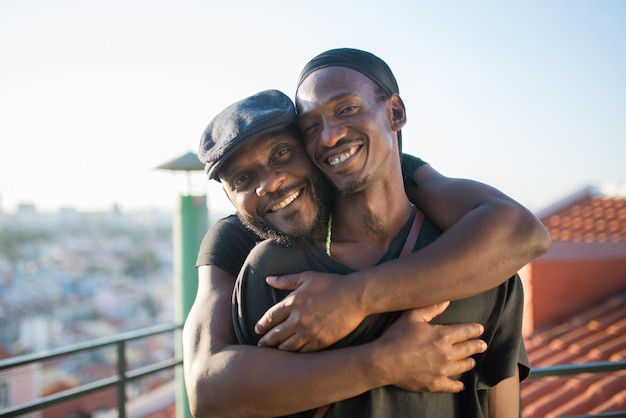 Primo piano di fidanzati gay africani felici che guardano l'obbiettivo. due uomini barbuti in abiti casual in piedi insieme sul tetto che si abbracciano per scattare foto sulla fotocamera del telefono. le persone lgbt amano, il concetto di felicità