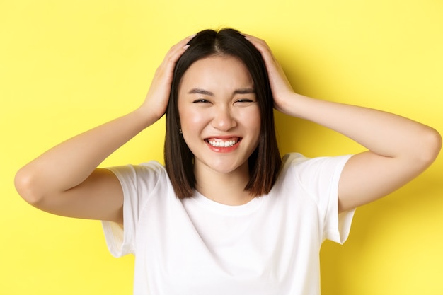 Primo piano di felice ragazza asiatica toccando i suoi capelli e sorridente, in piedi in maglietta bianca su giallo.