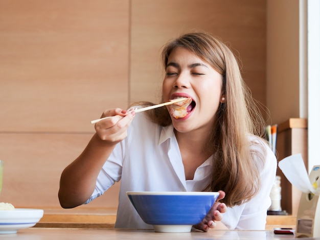 Закройте счастливое азиатское лицо женщины, используя палочки для еды в лапшу в ресторане, концепция времени обеда.