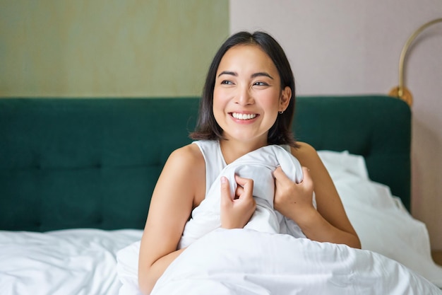 Close up of happy beautiful asian woman waking up in bed and\
enjoys morning looking outside window w