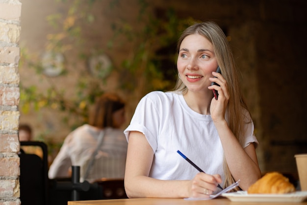 Close-up of a happy blonde woman in a white t-shirt looks out the window and talks on the phone, writing on paper while working at cafe at table with a croissant and a drink