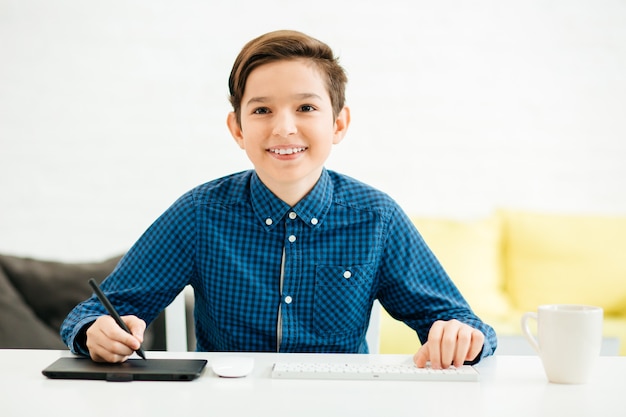 Close up of happy boy in checkered shirt sitting at the table and smiling while using a modern graphics tablet