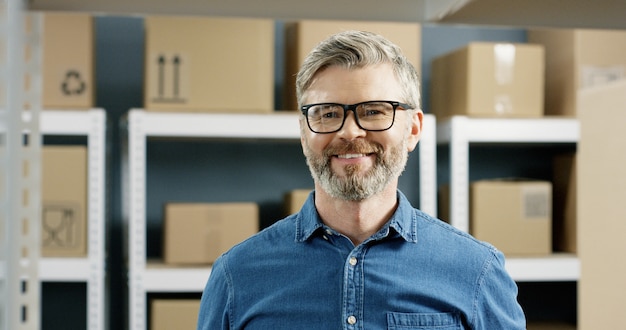 Close up happy smile postman in glasses standing in postal store with carton boxes on shelves. Post worker smiling joyfully in mail office among parcels.