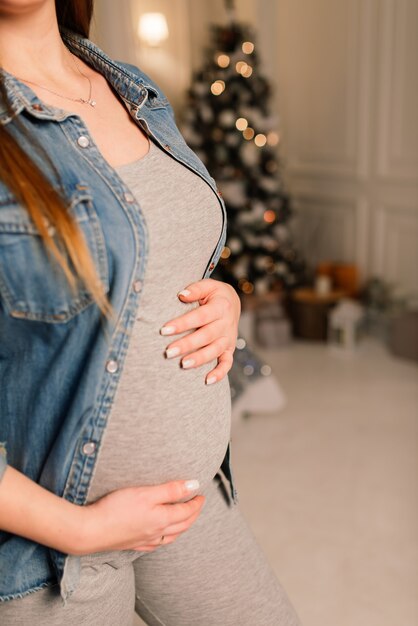 Close up of happy smiling pregnant woman touching her belly over christmas tree background