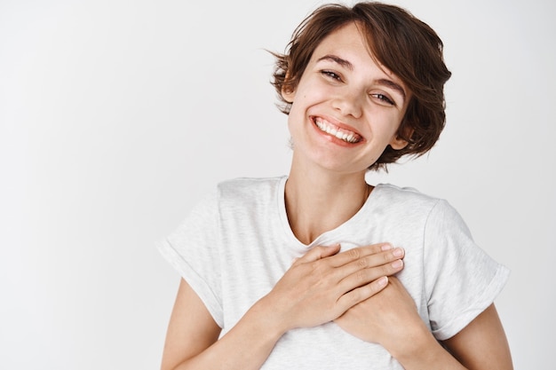 Primo piano di una donna sorridente felice dire grazie, tenendosi per mano sul cuore grato, esprimere gratitudine, in piedi contro il muro bianco