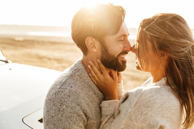 Primo piano di una giovane coppia felice innamorata che si abbraccia mentre si appoggia su un'auto sulla spiaggia soleggiata