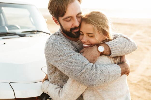 Close up of a happy young couple in love embracing while leaning on a car at the sunny beach