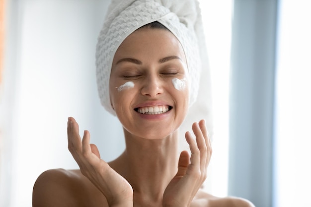close up head shot beautiful lady laughing while applying skincare moisturizing creme after morning showering happy young woman with wrapped in towel head enjoying smoothing skin domestic routine