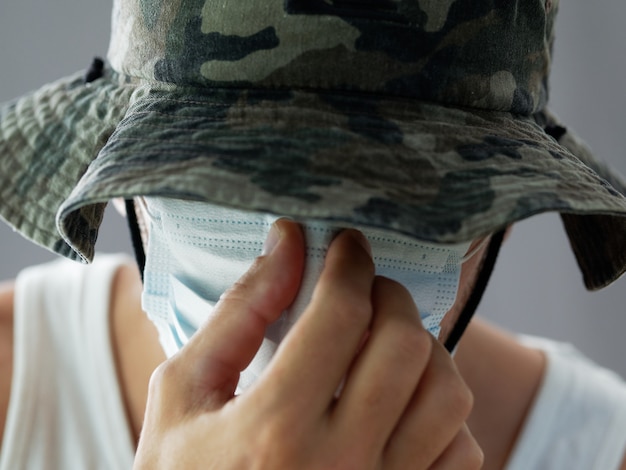 Close-up head shot. man in white t-shirt put medical mask.\
military style panama hat. gray background.