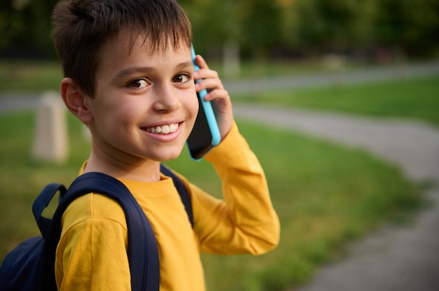 Close-up headshot portrait of a cheerful friendly adorable schoolboy, in yellow sweatshirt talking on mobile phone, smiling with toothy smile looking at camera on the city park background