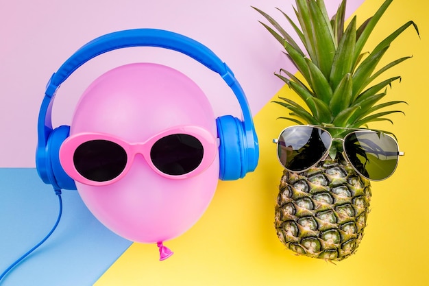 Close-up of helium balloon with sunglasses and headphones by pineapple over colored background