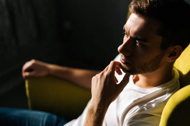 Close-up high-angle view face of serious handsome man thoughtfully touching chin searching solution sitting in yellow chair on dark background, studio shot, selective focus.