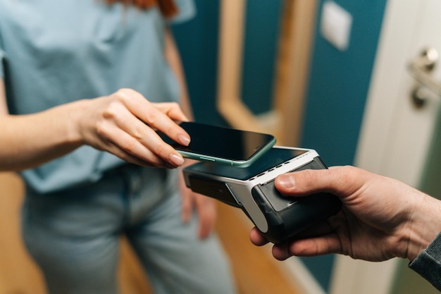 Foto close-up hoge hoekmening van onherkenbaar koeriersmannetje dat pos draadloze terminal geeft om contactloos te betalen met behulp van mobiele telefoon aan jonge vrouwelijke klant