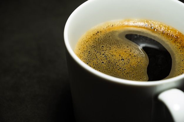 Close-up hot black coffee and bubbles in white cup against dark background