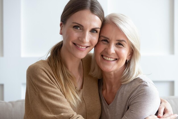 Close up image aged mother adult daughter sitting on couch\
indoors smiling looking posing for camera hugging feels happy spend\
time together concept of love relative people multigenerational\
family