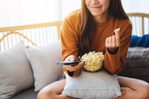 ポップコーンを食べて、自宅のソファに座ってテレビを見るためにリモコンでチャンネルを検索している美しい若い女性の画像をクローズアップ