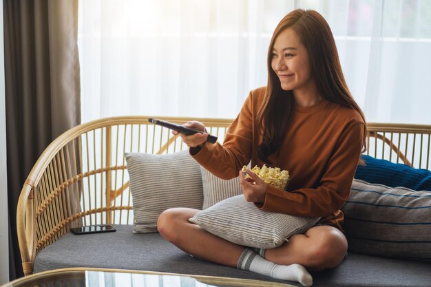 ポップコーンを食べて、自宅のソファに座ってテレビを見るためにリモコンでチャンネルを検索している美しい若い女性の画像をクローズアップ