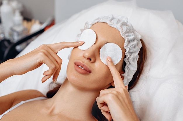 Close-up image in a beauty salon. A young girl is lying on a white couch. Holding cotton pads over her eyes. A white cap on her head. Protective safety measures before performing a cosmetic procedure.