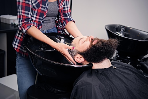 Close up image of female hairdresser washing bearded men's hair before haircut in a saloon.