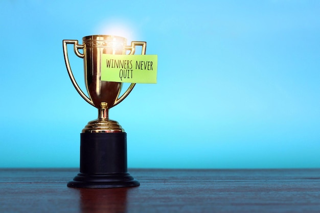 Close up image of gold cup trophy and sticky note with text WINNERS NEVER QUIT on blue background