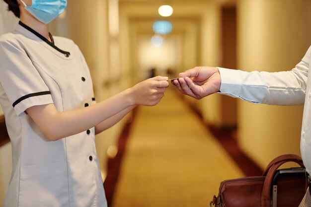 Photo close-up image of hotel manager in protective mask giving electronic key to man with big bag when they are standing in corridor