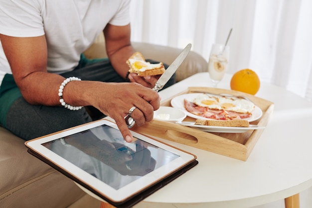 Photo close-up image of man eating breakfast and checking stock market data on scren of tablet computer