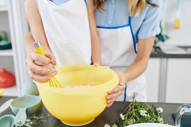 Close-up image of mother helping daughter to whisk eggs in big plastic bowl