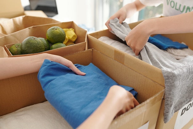 Close-up image of people packing tidy clothes and fresh food in cardboard boxes for people affected by natural disaster