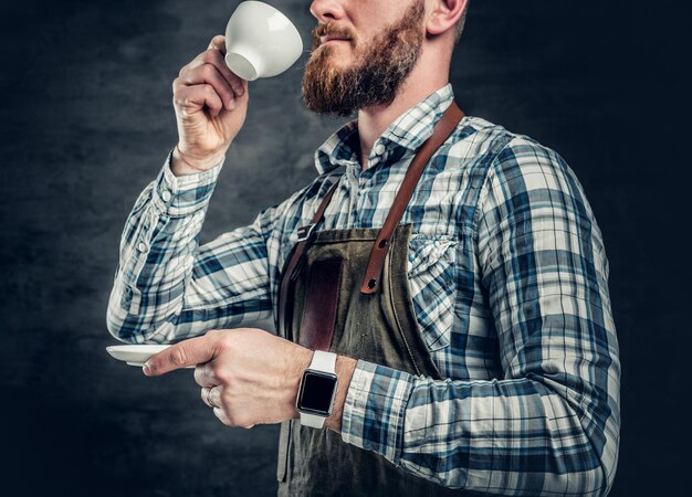 Close up image of redhead bearded male holds a cup of coffee over grey background.