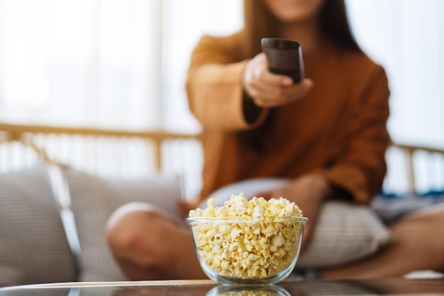 집에서 소파에 앉아 TV를 보기 위해 리모컨으로 팝콘을 먹고 채널을 검색하는 여성의 이미지를 닫습니다