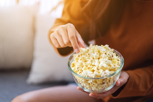 Close up image of a woman holding and picking pop corn from a bowl to eat while sitting on sofa at home