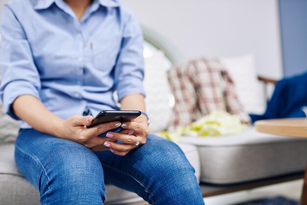 Close-up image of woman sitting on couch in messy room and texting friends instead of cleaning