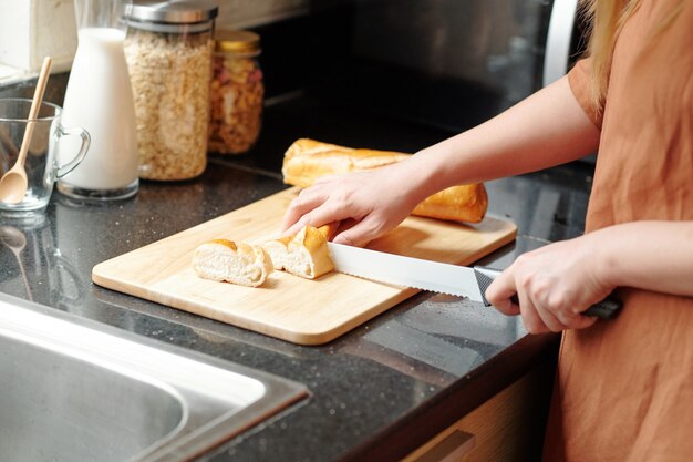Immagine ravvicinata di una giovane donna che taglia una baguette francese fresca su una tavola di legno al bancone della cucina