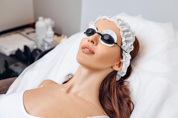 Close-up image. A young woman is lying on a white couch. White cotton pads and black safety glasses over her eyes. A white cap on her head. Protective safety measures before performing procedure.