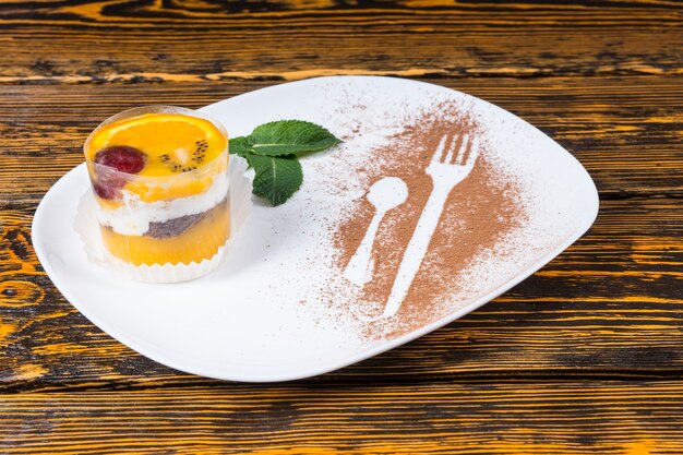 Close up di singoli decadenti torta di frutta dessert servita su un piatto bianco con contorno di foglie di menta e contorni di utensili spolverati di cacao sulla superficie del tavolo in legno